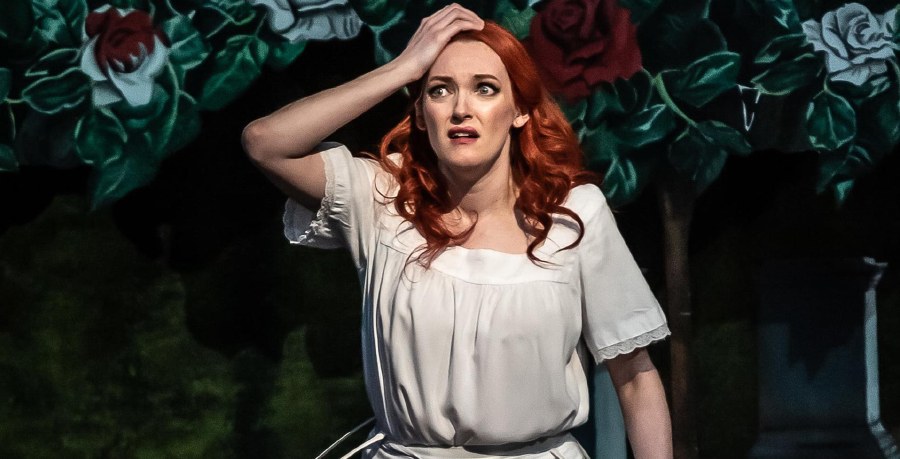 Soprano Jennifer France in a frilled white shirt holding her hand to her head in front of an opera scene of roses and greenery